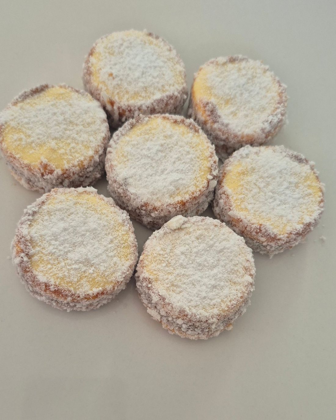 Siete alfajores de coco sobre una superficie clara, con azúcar glass arriba y el borde cubierto de coco rallado